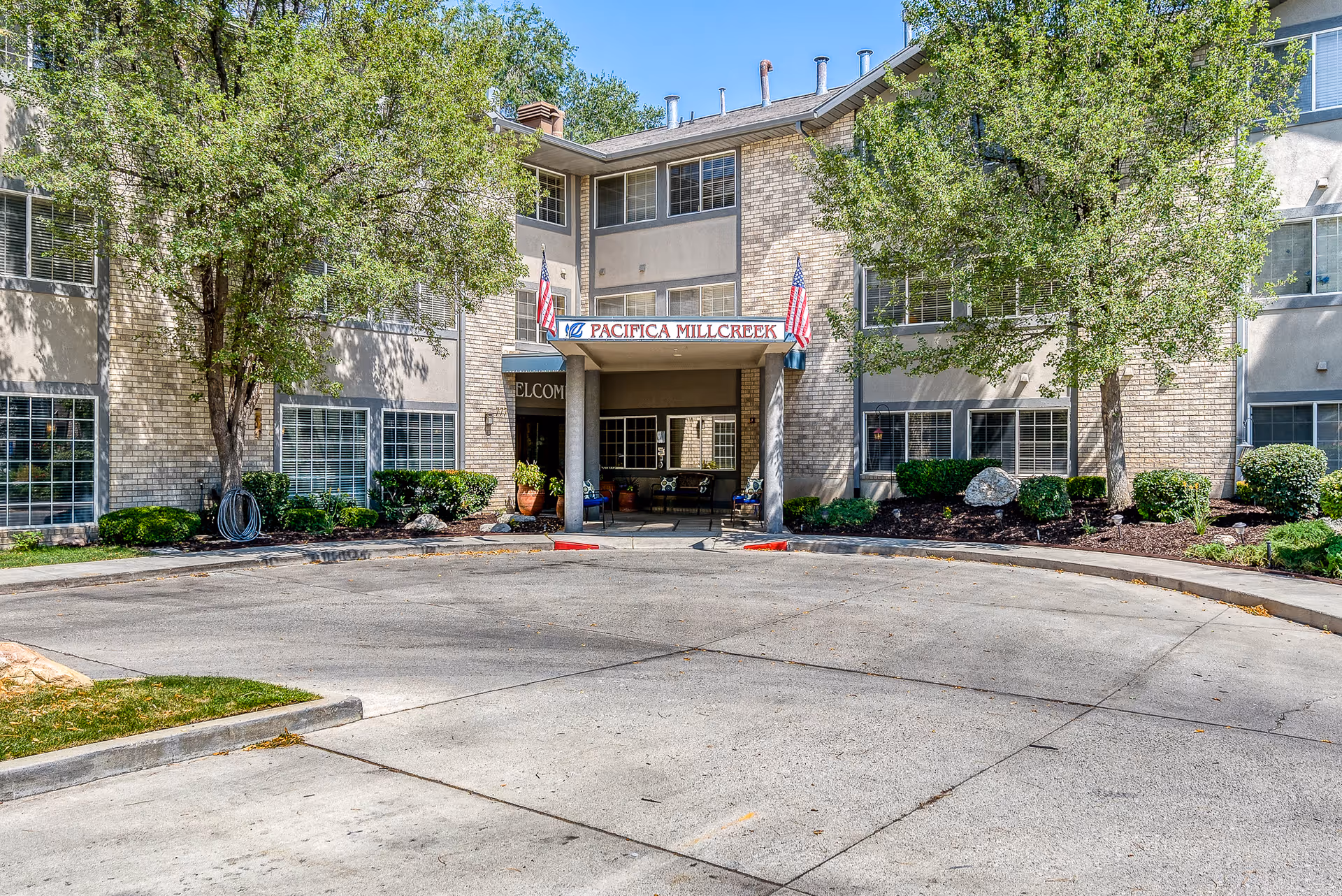 Front exterior view of a senior living facility named Pacifica Millcreek with a circular driveway, two American flags, trees, and shrubs around the entrance.