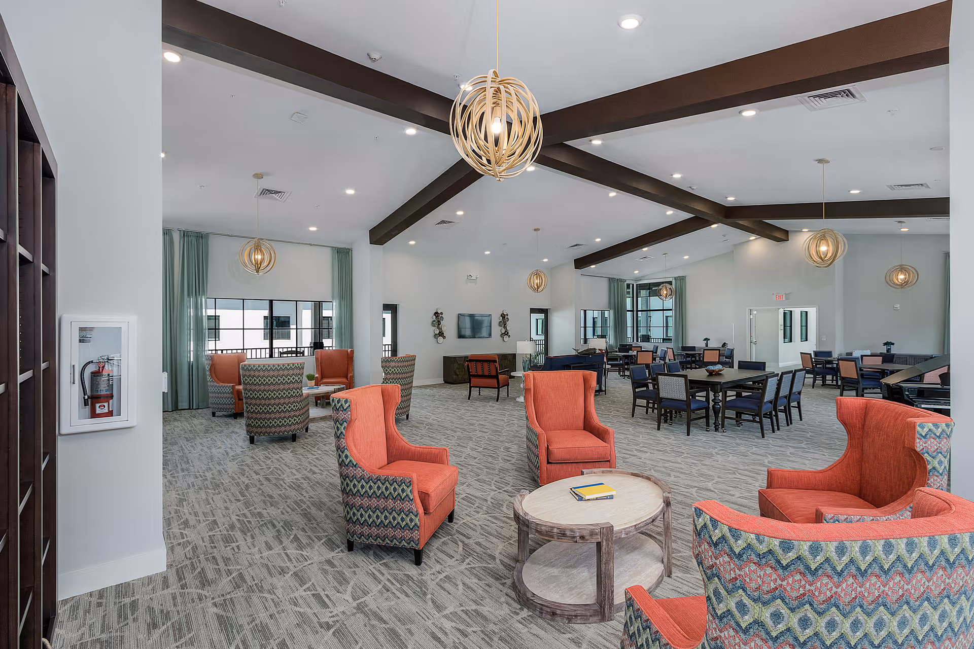 A spacious and well-lit common area in a senior living facility featuring multiple seating arrangements with colorful armchairs and tables. The room has a modern design with exposed dark ceiling beams, pendant lights, large windows with green curtains, and a television mounted on the far wall.