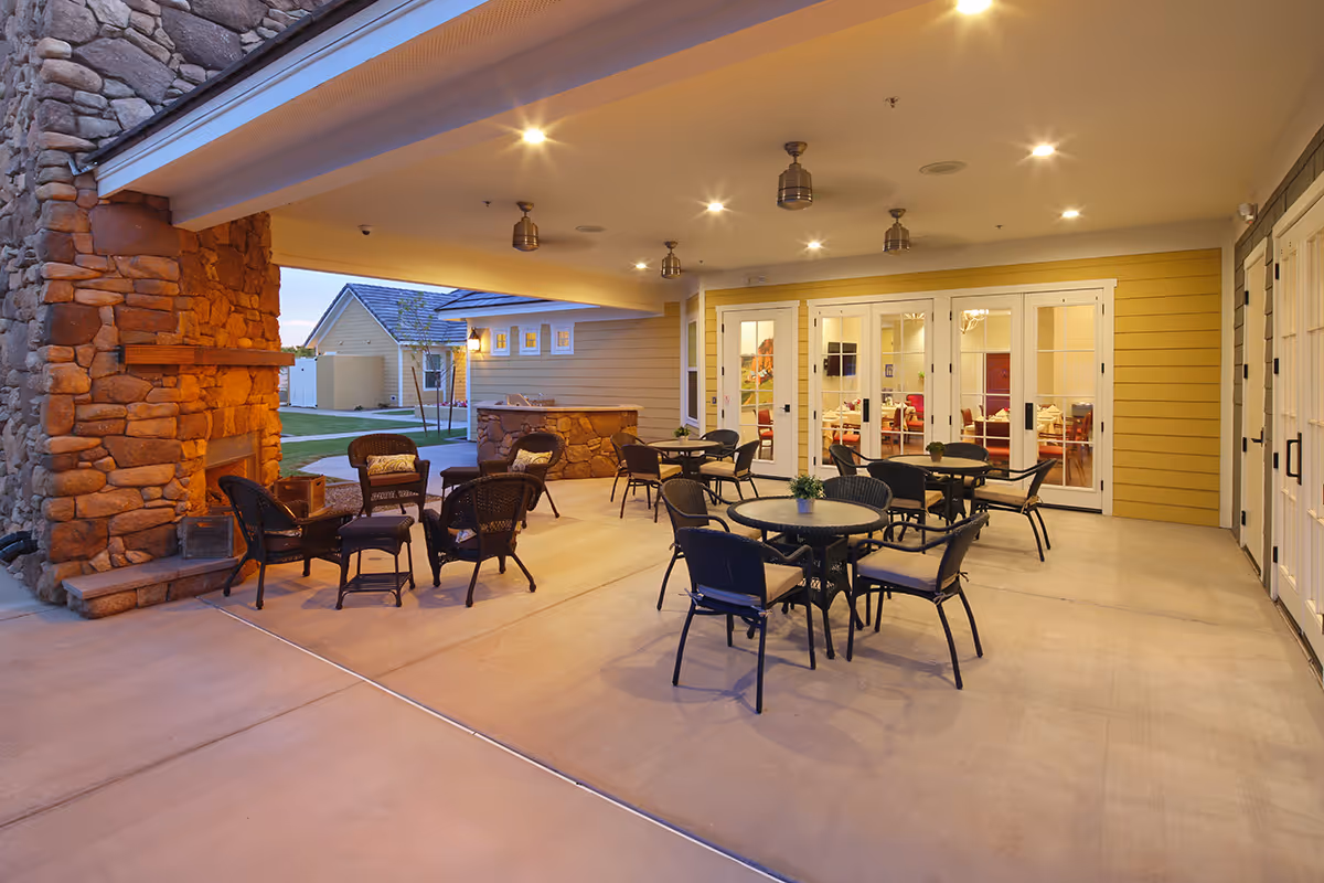Covered outdoor patio area with several round tables and chairs, a stone fireplace on the left, ceiling fans, and warm lighting. The patio is adjacent to a building with yellow siding and multiple glass doors leading inside.