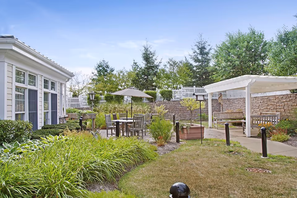 Outdoor patio area at Chapters Living of Barrington featuring a round table with chairs and an umbrella, surrounded by greenery and landscaping. There is a white pergola with benches underneath, a stone retaining wall, and a white fence in the background.