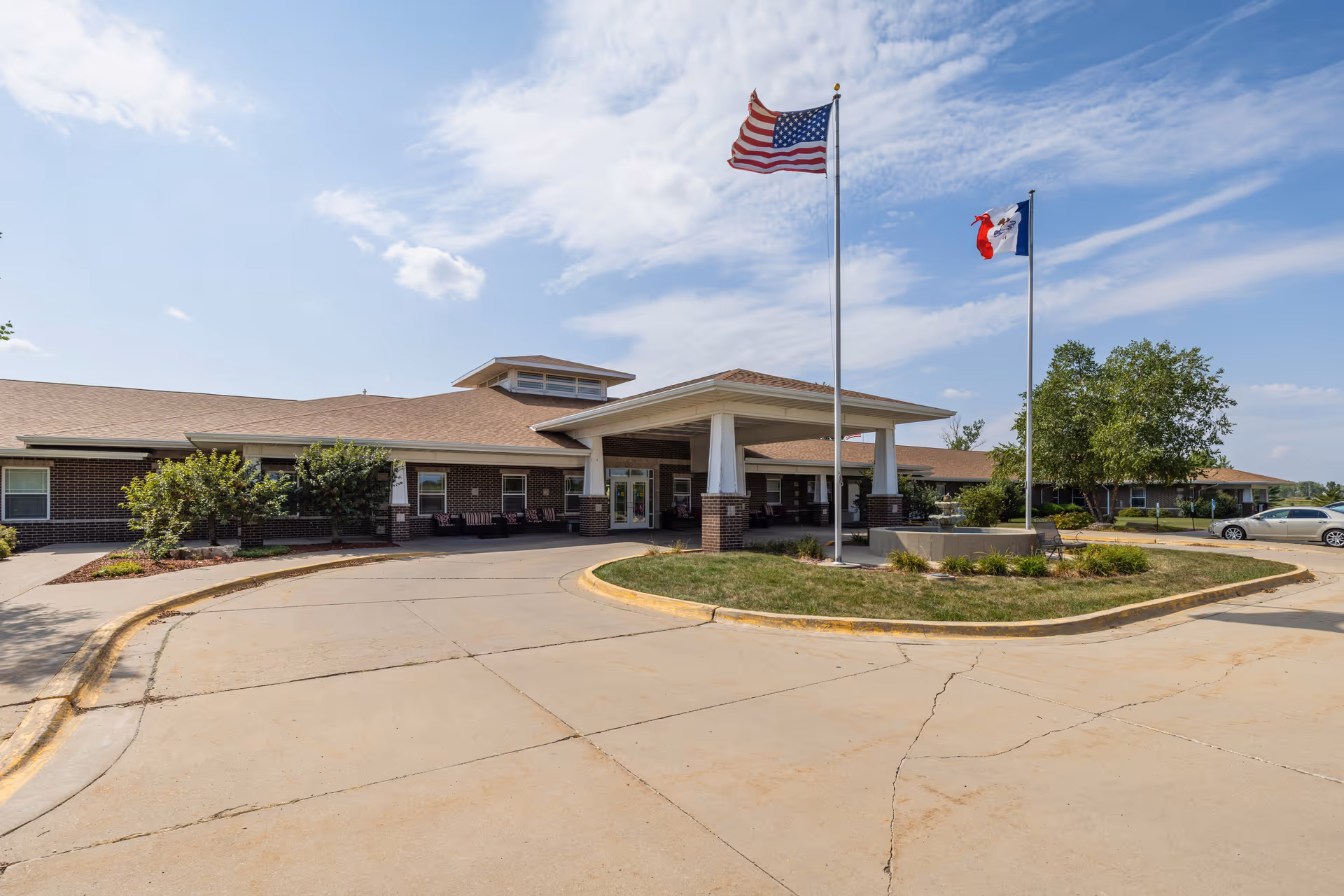 Exterior view of a single-story senior living facility building with a covered entrance and two flagpoles displaying the American flag and another flag. The building has a brick facade and a paved driveway with some landscaping and trees around it under a partly cloudy sky.
