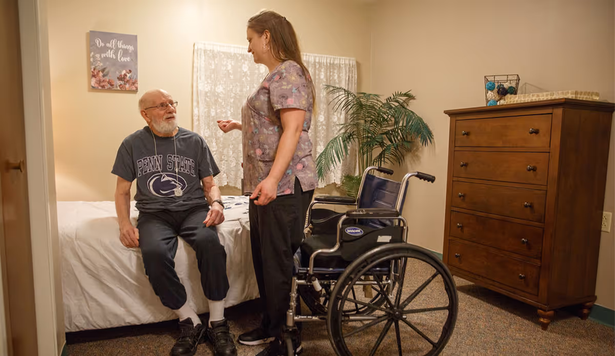 An elderly man wearing a Penn State t-shirt sits on the edge of a bed in a bedroom, talking with a female caregiver standing beside him. A wheelchair is positioned nearby, and the room contains a wooden dresser, a potted plant, and a wall decoration with the phrase 'Do all things with love.'