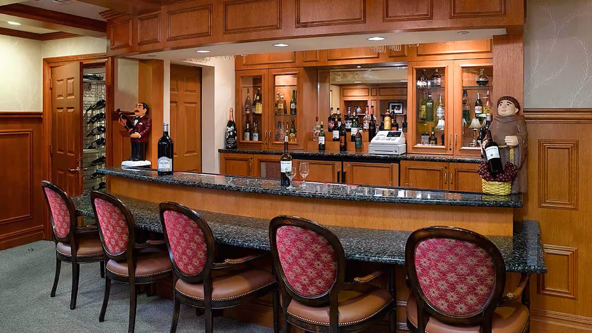 Interior view of a bar area with a granite countertop and six chairs with red patterned upholstery. Behind the bar are wooden cabinets with glass doors displaying various bottles of alcohol. Two decorative figurines holding wine bottles are placed on the bar counter. The walls have wood paneling and light-colored wallpaper.