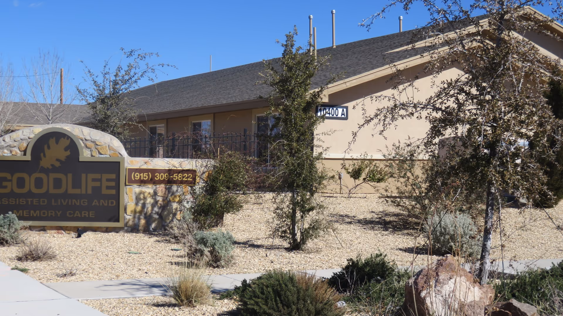 Exterior view of Good Life Assisted Living and Memory Care El Paso facility showing a stone sign with the facility name and phone number, surrounded by desert landscaping with small trees and bushes, and a beige building in the background.