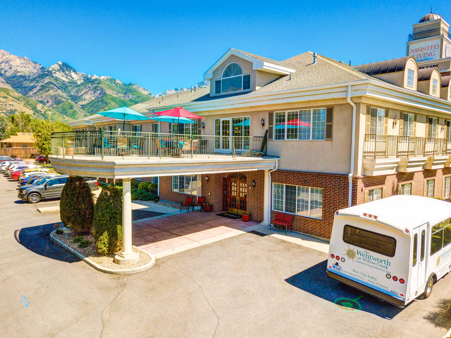 Front exterior of the Escalante at Willow Creek assisted living building showing a covered entrance, second-story terrace with umbrellas, parked shuttle bus, and mountains in the background.