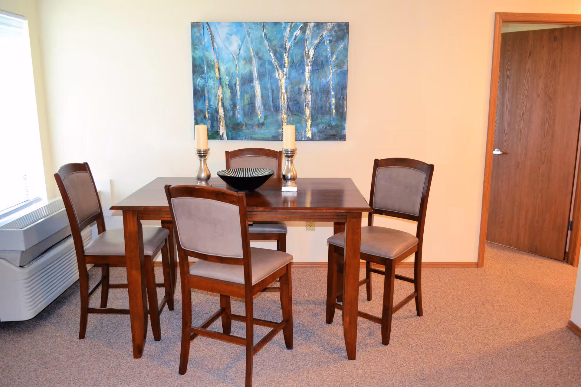 A dining area with a wooden table and four matching chairs with cushioned seats. On the table are two silver candle holders with cream-colored candles and a decorative bowl. A large painting of birch trees with blue and green tones hangs on the wall behind the table. There is a window with blinds on the left side and a wooden door on the right side of the room.