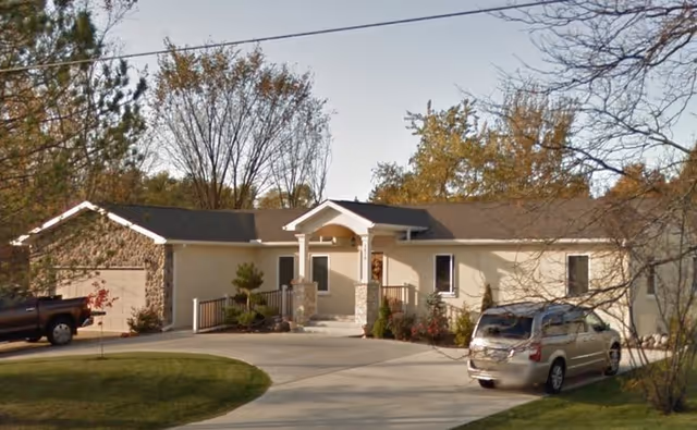 Single-story residential building with a stone and beige exterior, a covered front porch with columns, surrounded by trees and a curved driveway with two parked vehicles.