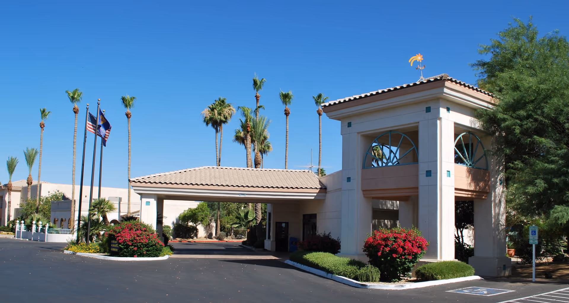 Exterior view of the entrance to a senior living facility with a covered drop-off area, surrounded by palm trees and landscaped bushes under a clear blue sky.