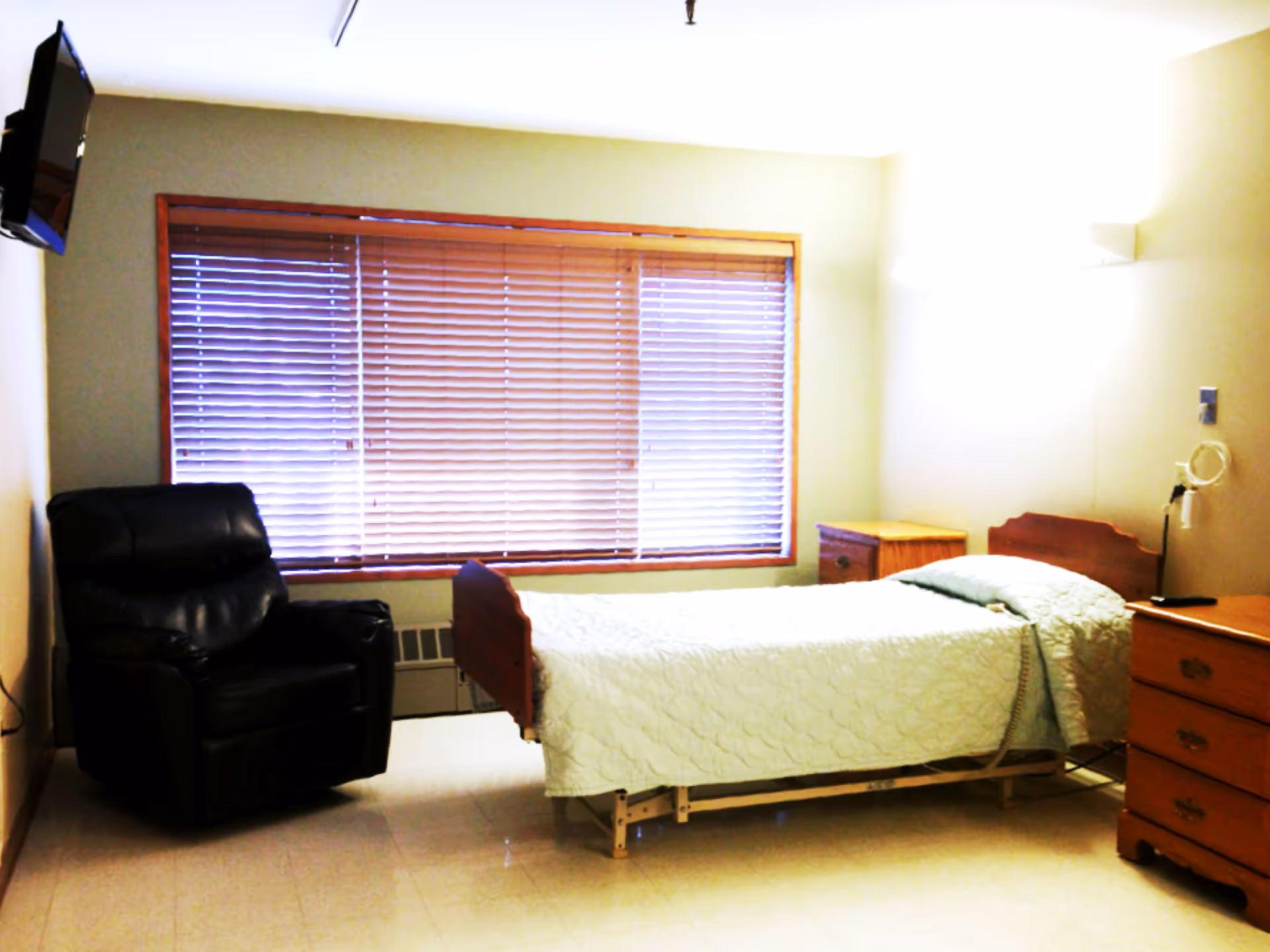 A simple bedroom with a single bed covered with a light green quilt, a black recliner chair, a wooden dresser, a wooden nightstand, and a large window with closed blinds. A wall-mounted TV is visible in the upper left corner.