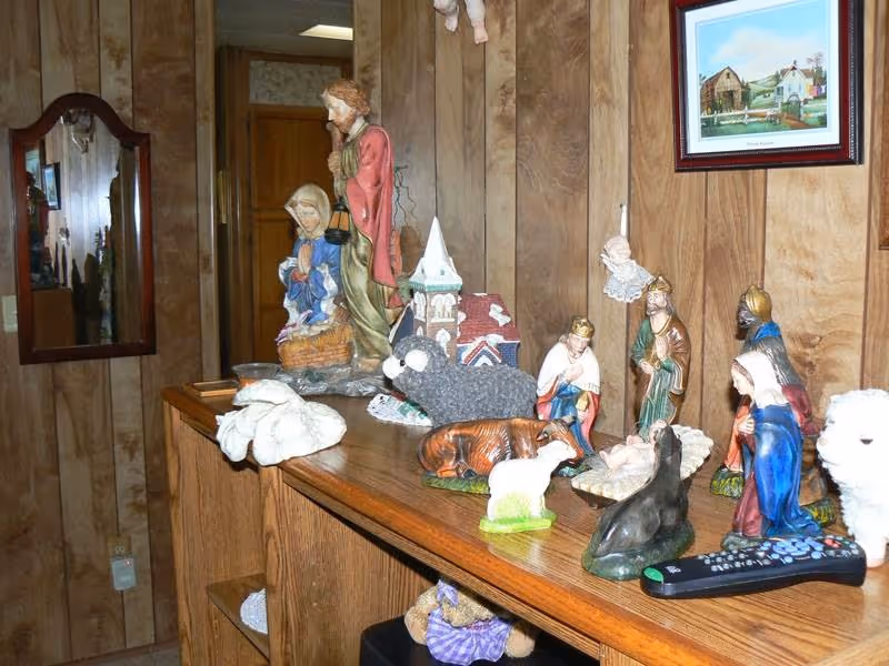 A wooden shelf with various nativity scene figurines including Mary, Joseph, baby Jesus, the three wise men, animals, and an angel. The background shows wood-paneled walls, a mirror, and a framed painting of a barn and countryside.