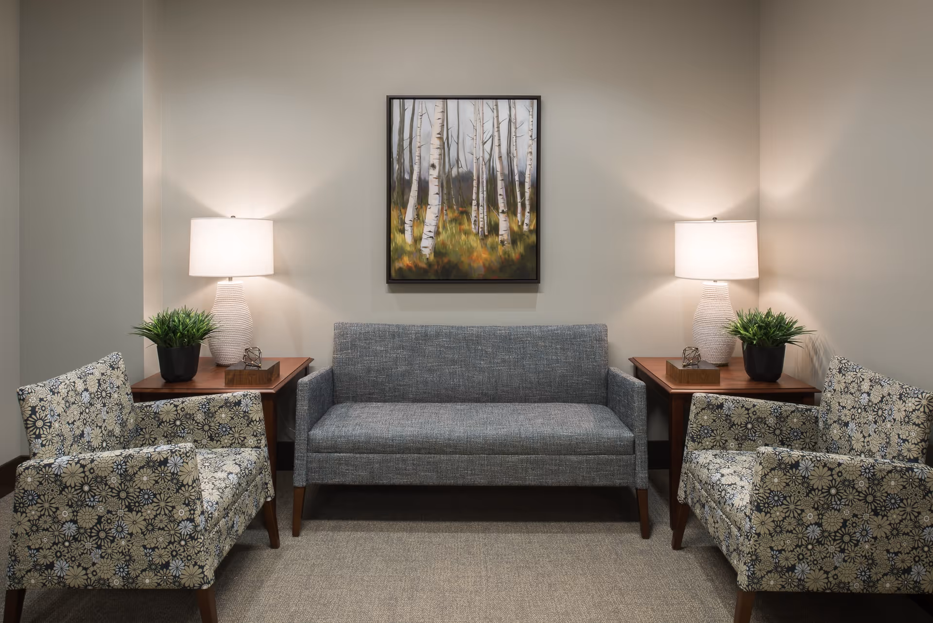 A cozy sitting area with a gray upholstered sofa centered between two patterned armchairs. Each armchair is positioned next to a wooden side table with a white lamp and a small green plant. A framed painting of birch trees hangs on the beige wall above the sofa.