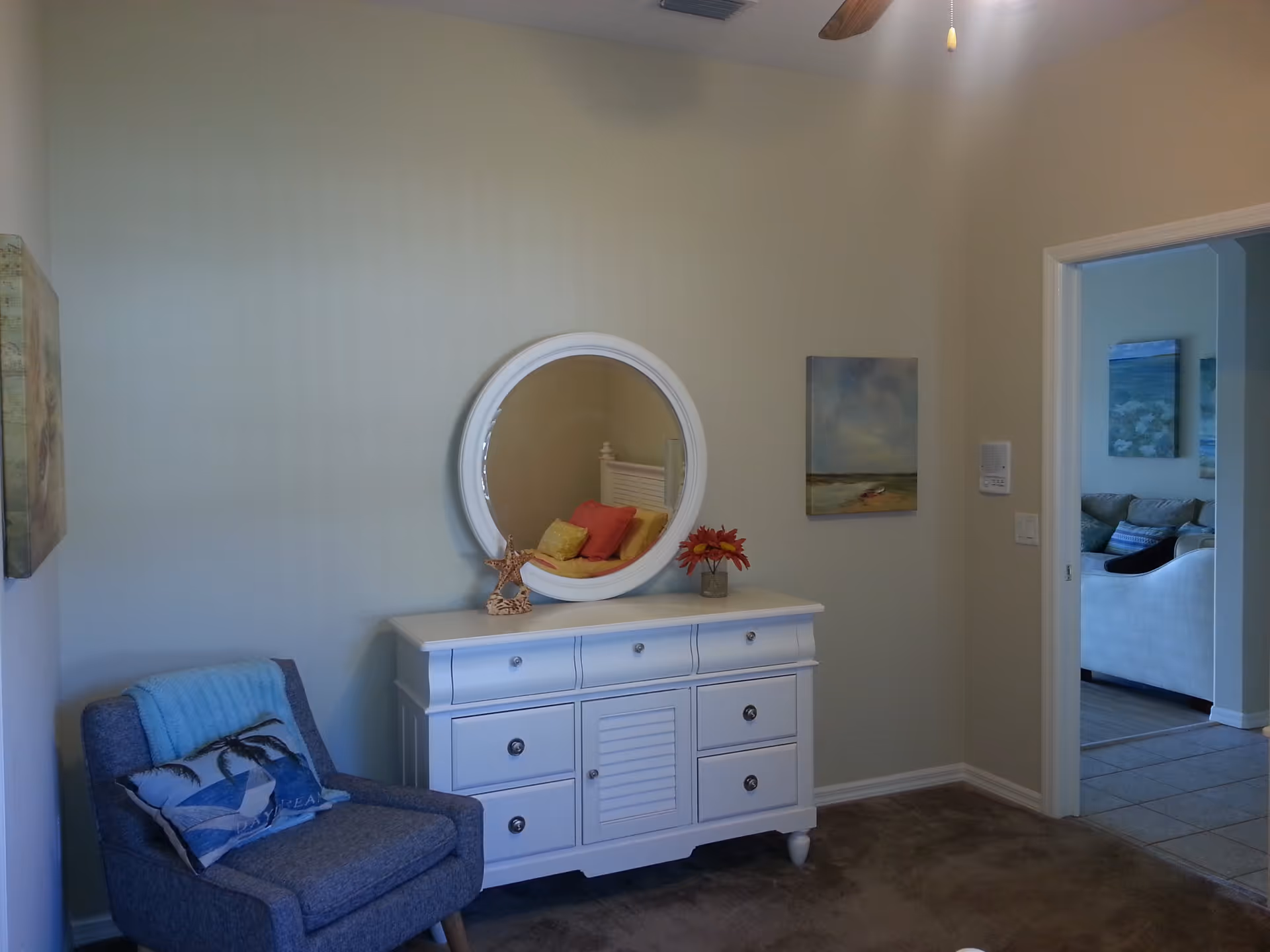 Interior sitting area with a white dresser topped by a round mirror and flowers, a gray armchair with pillows, and a doorway leading to a living room.
