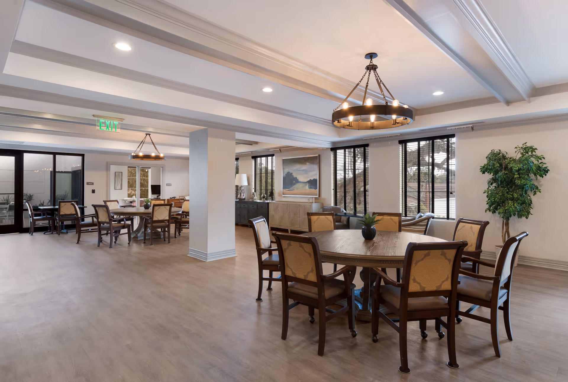 A spacious, well-lit common area with multiple round wooden tables surrounded by cushioned chairs. The room features large windows with black blinds, a potted plant, a sideboard with decorative items, and modern ceiling light fixtures. The floor is light wood, and there is an exit sign visible in the background.