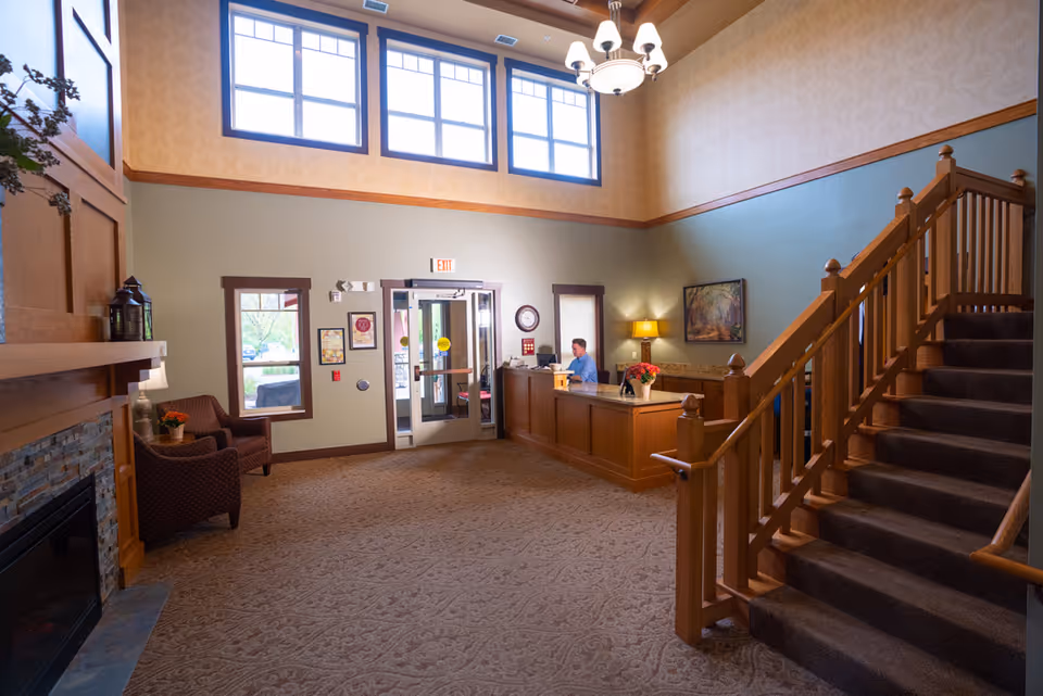 Bright assisted living lobby with a reception desk, carpeted seating area, wooden staircase, and large upper windows.