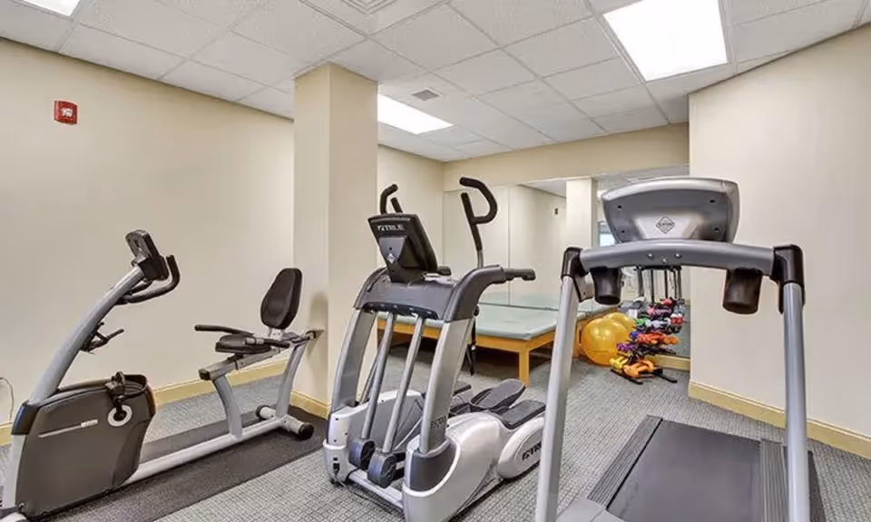 A small fitness room with exercise equipment including a recumbent bike, elliptical machine, and treadmill. The room has beige walls, a carpeted floor, a large mirror on one wall, and some exercise balls and weights in the corner.