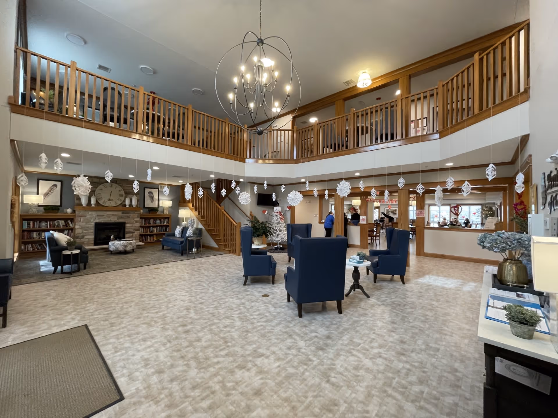 Spacious senior living facility common area with high ceilings and a large modern chandelier. The room features a seating area with blue armchairs and small tables in the center, a fireplace with a clock and decorative items on the left, and a reception desk on the right. There are hanging white paper snowflake decorations from the ceiling. A wooden staircase leads to an upper balcony overlooking the area.