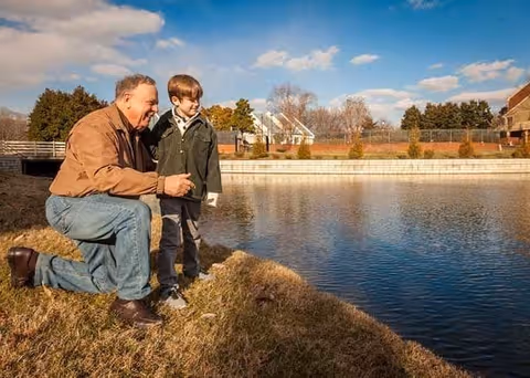 An older man kneeling on the grass beside a young boy near a calm body of water with houses and trees in the background under a partly cloudy sky.