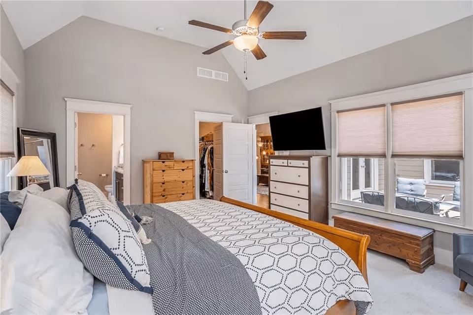 A bedroom with a large bed covered in patterned bedding and multiple pillows. The room has a ceiling fan with a light, two dressers, a wall-mounted flat-screen TV, and three windows with blinds partially closed. There is a door leading to a walk-in closet and another door leading to a bathroom. A wooden chest and a gray armchair are placed near the windows.