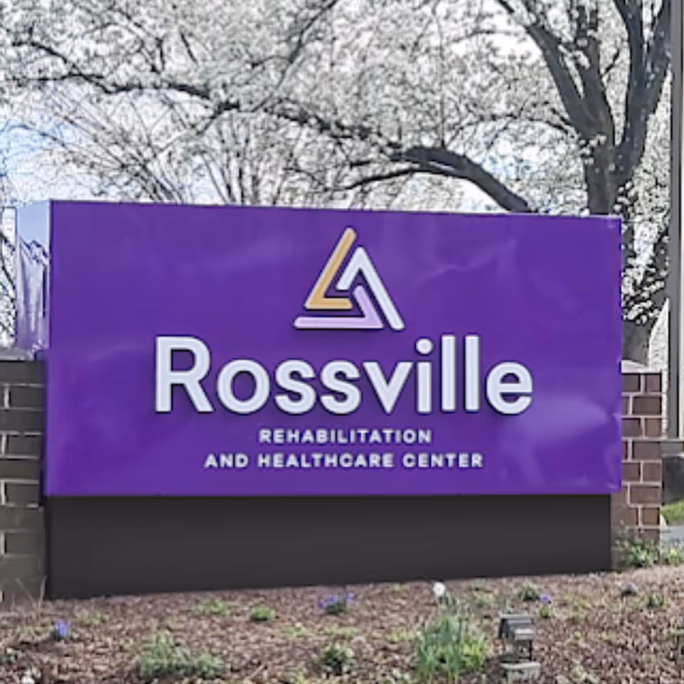 A large purple sign with the logo and text 'Rossville Rehabilitation and Healthcare Center' situated outdoors with trees and a brick base.