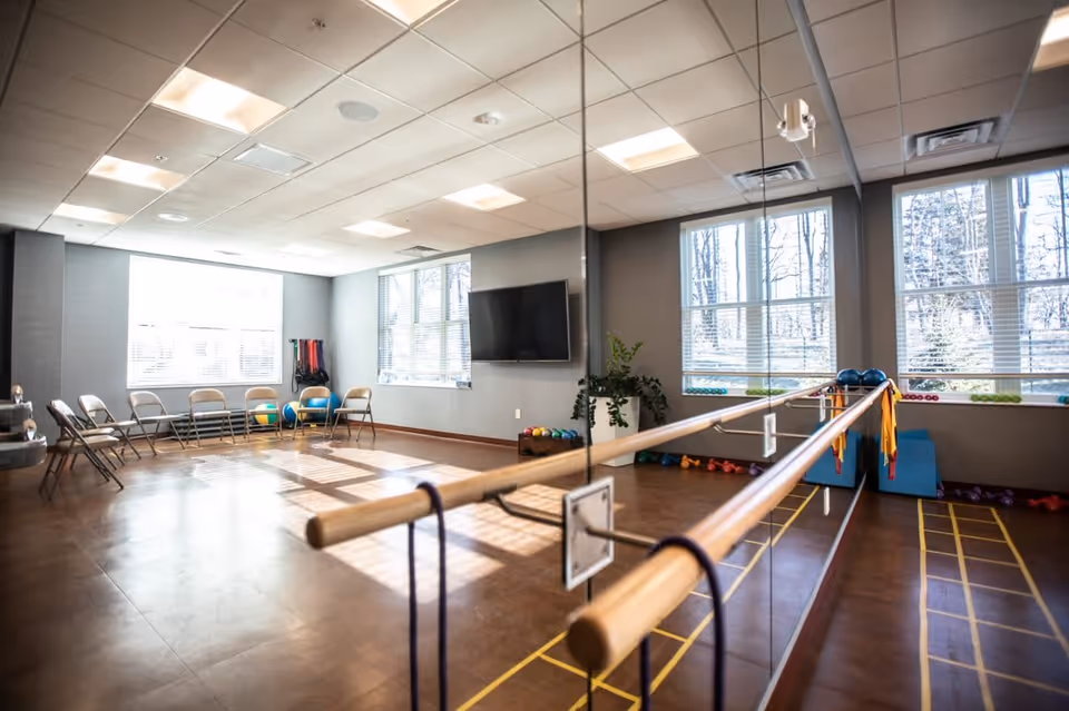Bright exercise room with large windows letting in natural light, a wall-mounted TV, wooden ballet barre with resistance bands, folding chairs arranged along the wall, and various exercise equipment including medicine balls and dumbbells.