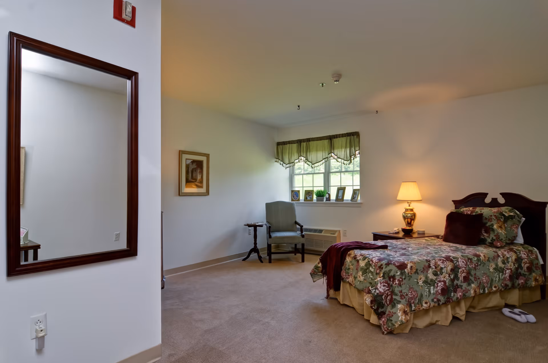 A senior living bedroom with a single bed covered in a floral bedspread, a bedside table with a lamp, a cushioned armchair next to a window with green valance curtains, a small side table, and a large wall mirror on the left side.