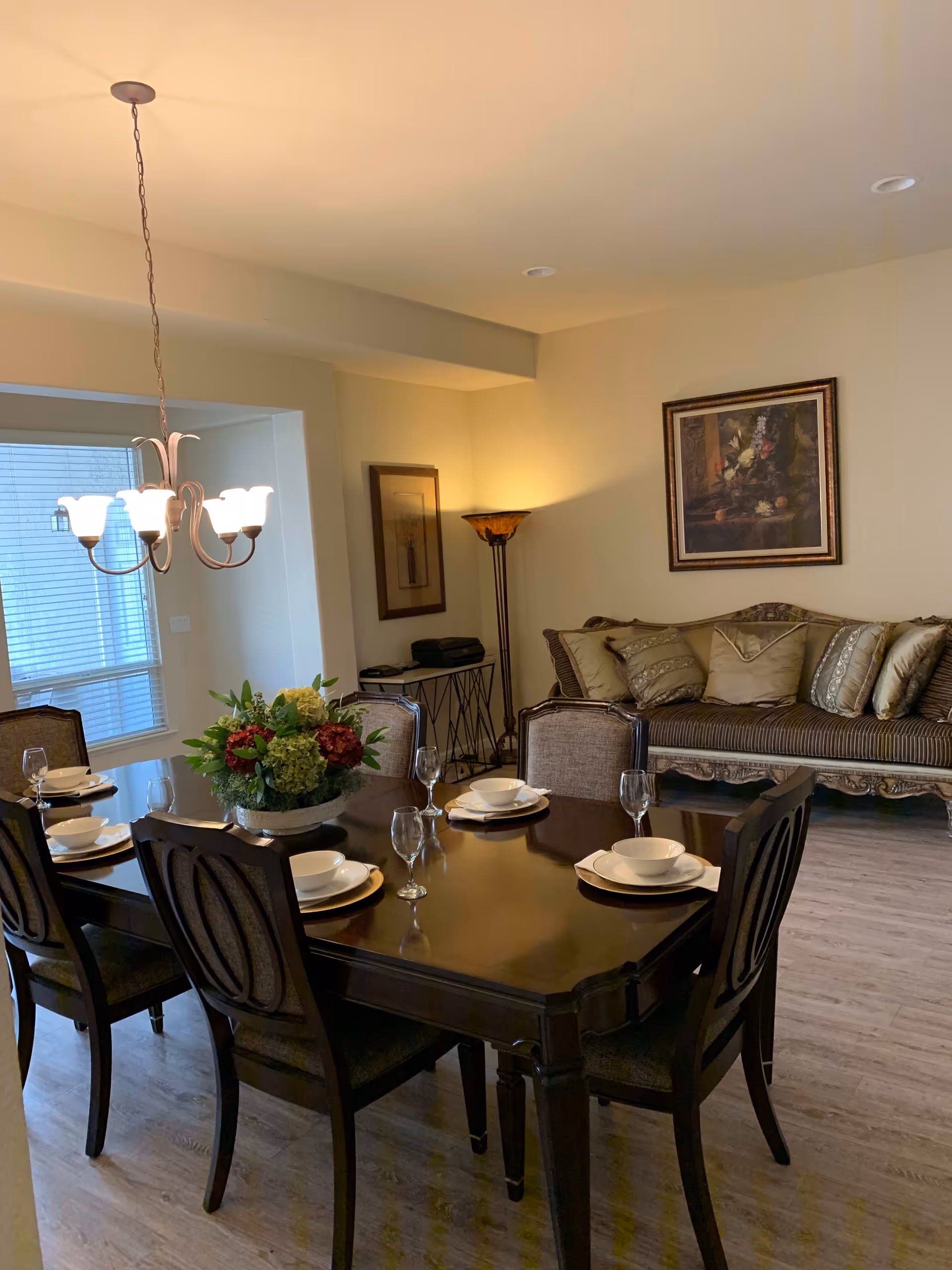 A dining room with a dark wooden dining table set for six people with white plates, bowls, and wine glasses. A floral centerpiece is on the table. Behind the dining area is a sitting area with a cushioned sofa adorned with multiple pillows. The room has light-colored walls, a floor lamp in the corner, framed artwork on the walls, and a chandelier hanging above the dining table. A window with blinds is visible on the left side.