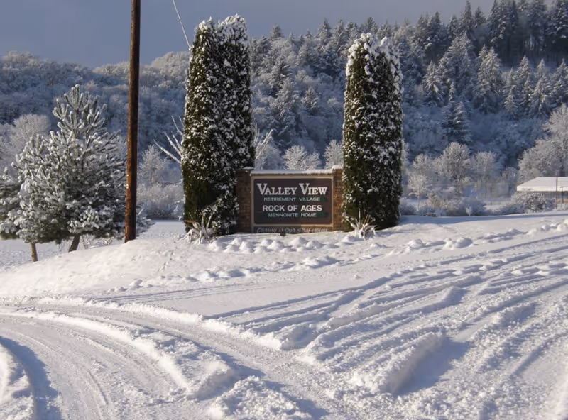 Snow-covered landscape with tire tracks in the snow leading to a sign that reads 'Valley View Retirement Village Rock of Ages Mennonite Home' flanked by two tall snow-dusted evergreen trees, with a forested hill in the background.