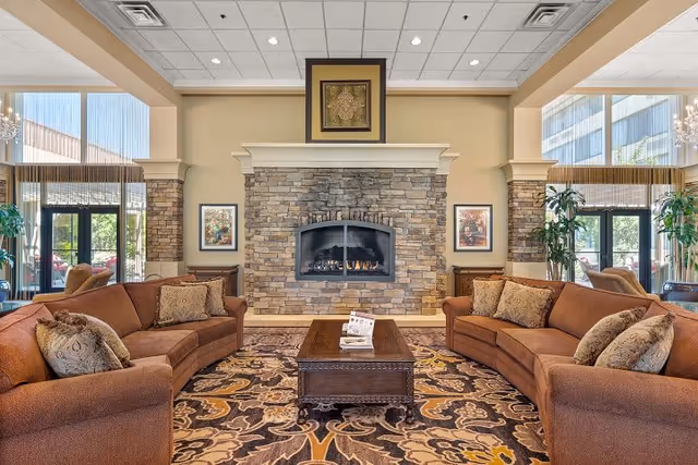 A cozy living room area with two brown sofas facing each other across a wooden coffee table, centered in front of a stone fireplace with a fire burning. The room features large windows with natural light, decorative pillows on the sofas, framed artwork on the walls, and a patterned area rug covering the floor.