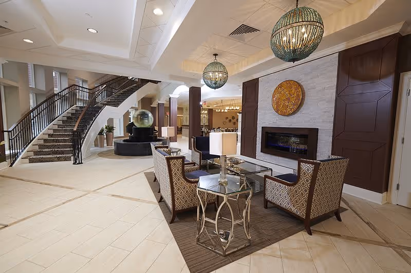 A spacious and elegant senior living facility common area with a seating arrangement consisting of four patterned armchairs around a glass coffee table on a brown rug. The area features a modern fireplace set in a white stone wall with decorative art above it. Two large, spherical pendant lights hang from the ceiling. In the background, there is a curved staircase with a dark railing and a large water fountain with a glass sphere. The floor is tiled in a light color, and the space is well-lit with natural and artificial lighting.