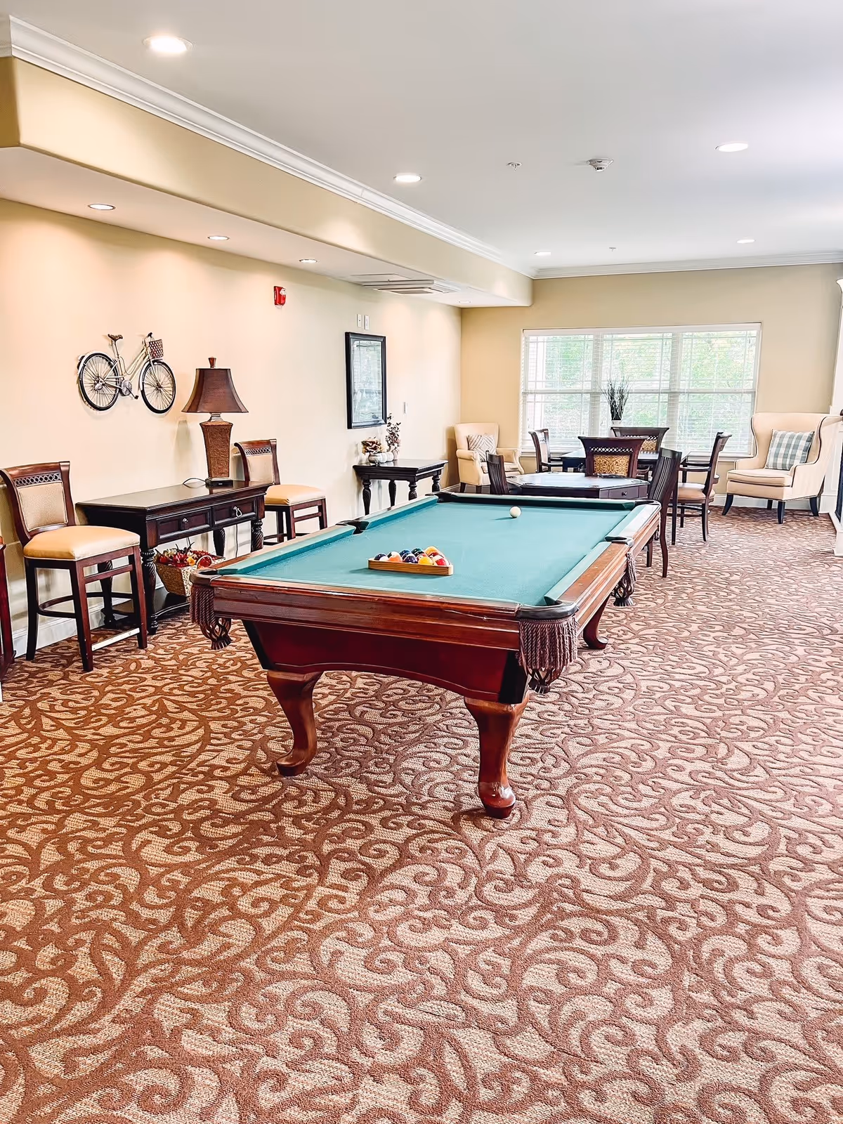 A well-lit recreational room with a pool table in the center, set up with billiard balls and a cue ball. The room features patterned carpet flooring, beige walls, and recessed ceiling lights. Along one wall, there are chairs, a table with a lamp, and decorative items including a wall-mounted bicycle sculpture. In the background, there are several tables and chairs near large windows with natural light, and comfortable armchairs with cushions.