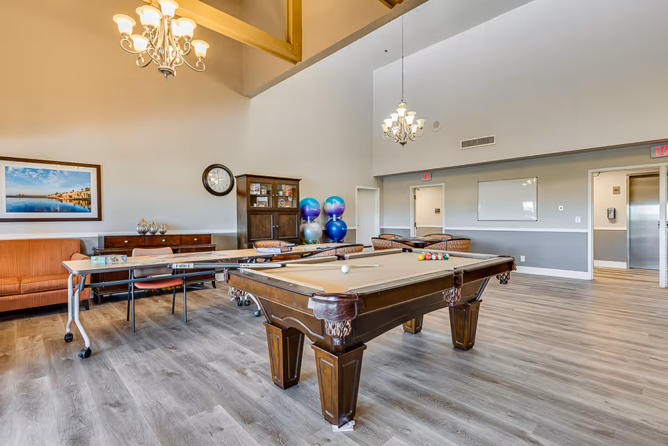 Spacious activity room with a pool table, tables and chairs, a couch, and stacked exercise balls under chandeliers.