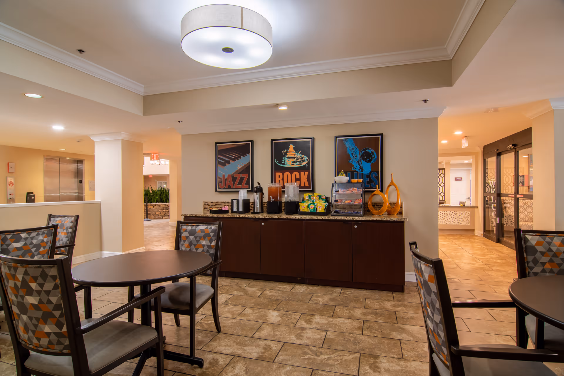 A common area in Dunwoody Pines Retirement Community featuring round tables with patterned chairs, a countertop with beverage dispensers, snacks, and decorative items. Three framed posters with the words Jazz, Rock, and Blues hang on the wall above the countertop. The space has tiled flooring and warm lighting.