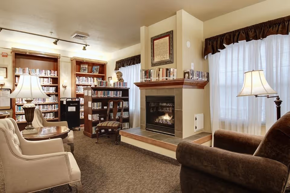 A cozy library room with bookshelves filled with books, a fireplace with a framed document above it, and comfortable seating including armchairs and a wooden chair. The room is softly lit by table lamps and natural light coming through sheer curtains on the windows.