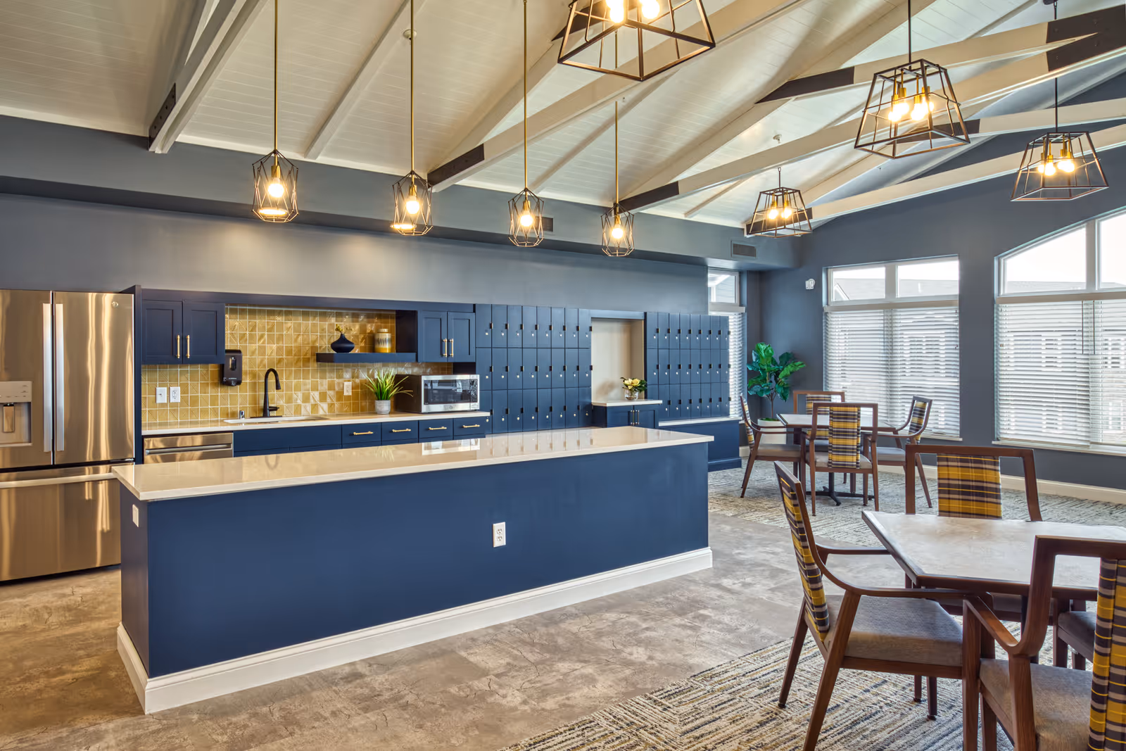 A modern kitchen and dining area in a senior living facility with blue cabinetry, a large island with a white countertop, stainless steel refrigerator, and multiple pendant lights hanging from a vaulted ceiling. There are several tables and chairs with plaid cushions near large windows letting in natural light.
