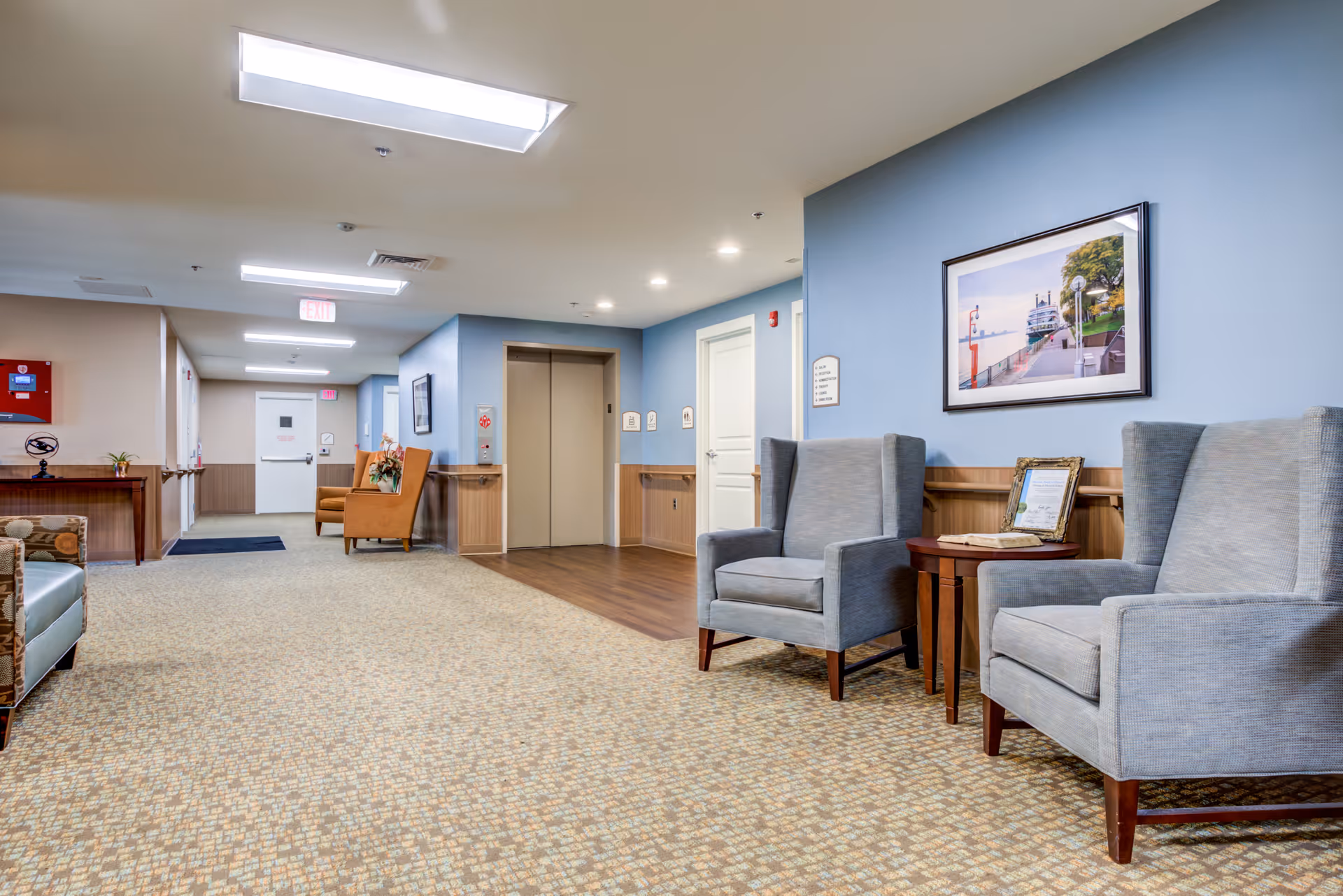 A well-lit hallway in a nursing and physical rehabilitation center featuring a seating area with two gray armchairs and a small wooden table. The walls are painted blue with wood paneling, and there is a framed picture hanging above the chairs. The hallway has carpeted flooring with a patterned design and leads to an elevator and several doors. Additional seating with orange chairs is visible further down the hallway.