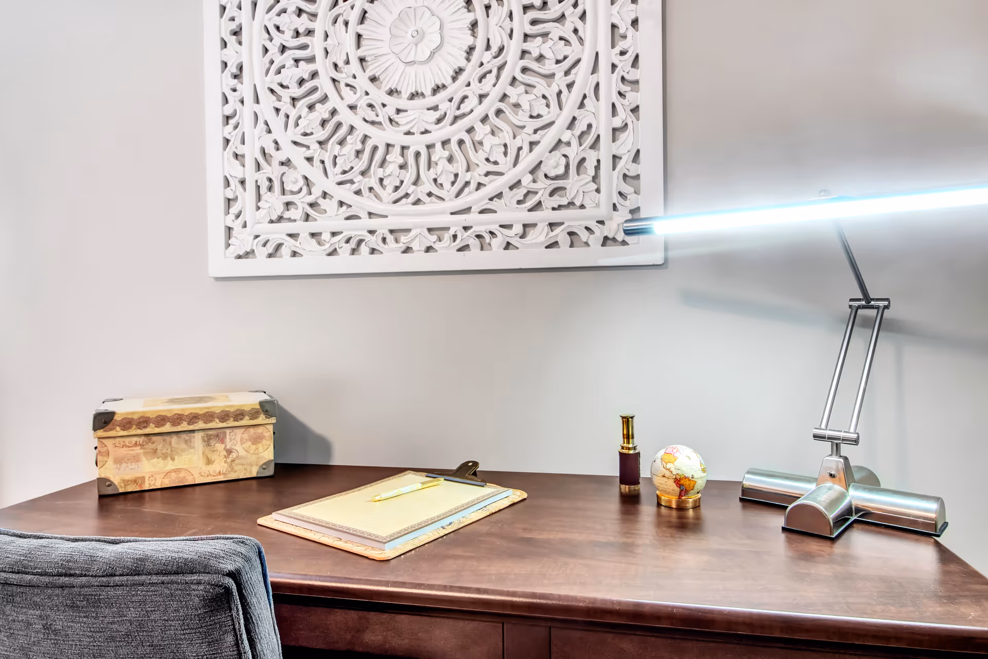 A wooden desk with a gray upholstered chair in front of it. On the desk are a decorative box, a clipboard with a notepad and pen, a small globe, a small telescope, and a modern silver desk lamp. Above the desk is a white, intricately carved wall decoration.