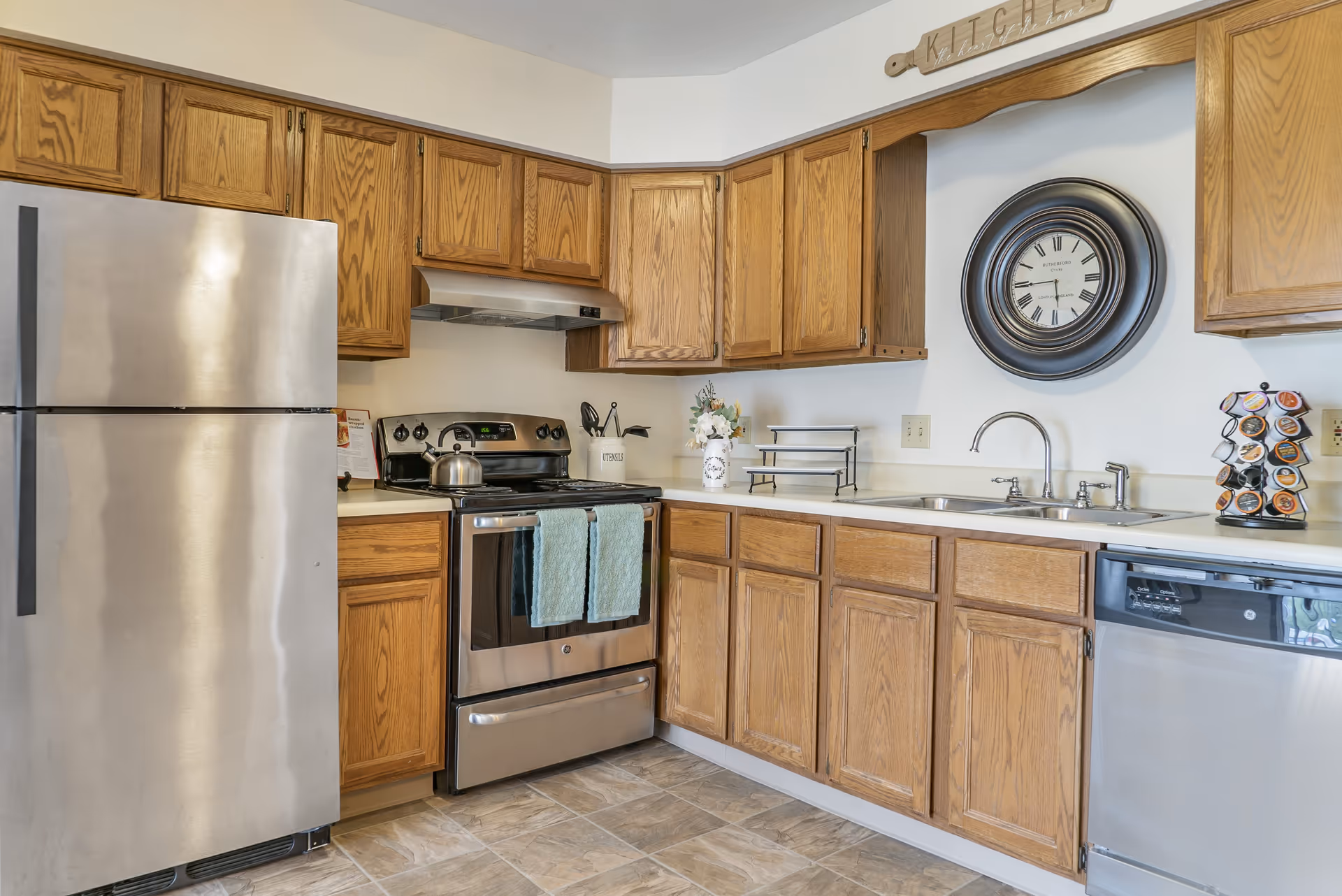 A kitchen with wooden cabinets, a stainless steel refrigerator, stove with two teal towels hanging on the handle, a double sink, dishwasher, and a wall clock above the sink. There is a small rack with coffee pods and a decorative sign above the clock that reads 'Kitchen the heart of the home.'