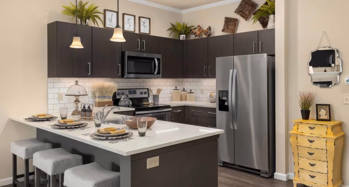 Modern kitchen with dark wood cabinets, stainless steel refrigerator and stove, white subway tile backsplash, and a white countertop island with three gray cushioned stools. The island is set with plates, bowls, glasses, and utensils. Decorative plants and framed artwork are placed on top of the cabinets and a small yellow chest with a plant and framed photo is against the wall.