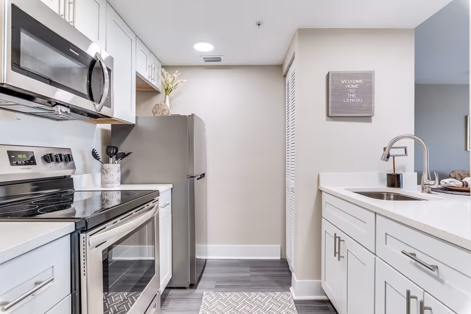 Modern kitchen with white cabinets, stainless steel appliances including a microwave, stove, and refrigerator. A countertop with a sink and faucet is visible on the right side. A small decorative sign on the wall reads 'WELCOME HOME TO THE LENOX!'. The floor has a gray patterned rug and dark wood flooring.