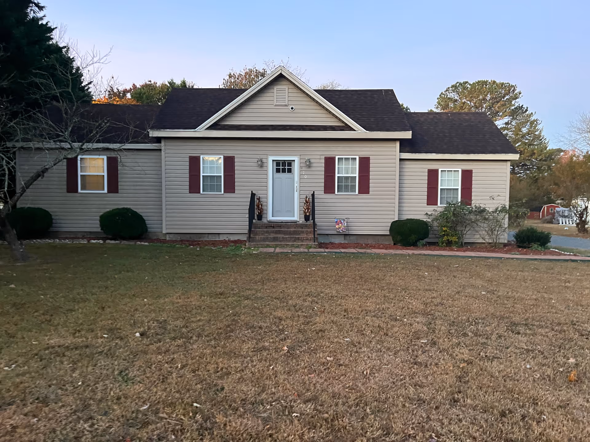 Front exterior view of a single-story house with beige siding, a dark shingled roof, and maroon shutters on the windows. There is a small set of stairs leading to a white front door with two black railings. The house is surrounded by a lawn with some bushes and trees, and the sky is clear.