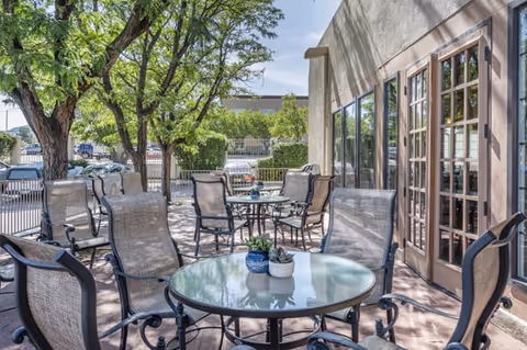 Outdoor patio area with multiple round glass tables and metal chairs with mesh backs and seats. The patio is shaded by trees and adjacent to a building with large windows and French doors. There are potted plants on the tables and a parking lot visible in the background.
