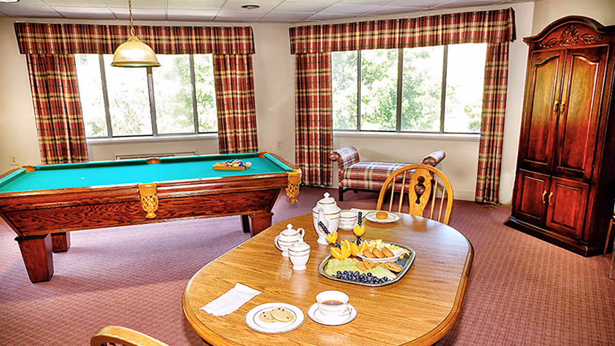 A bright room with a pool table, a wooden dining table set with tea and snacks, a plaid upholstered bench near large windows with plaid curtains, and a wooden armoire in the corner.