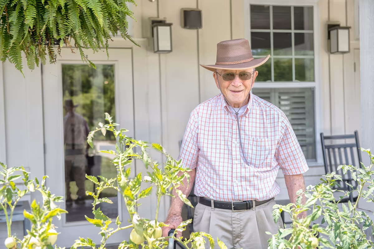 An elderly man wearing a brown hat, sunglasses, a checkered short-sleeve shirt, and beige pants stands outdoors in front of a white building with a glass door and window. He is surrounded by green plants and appears to be smiling.