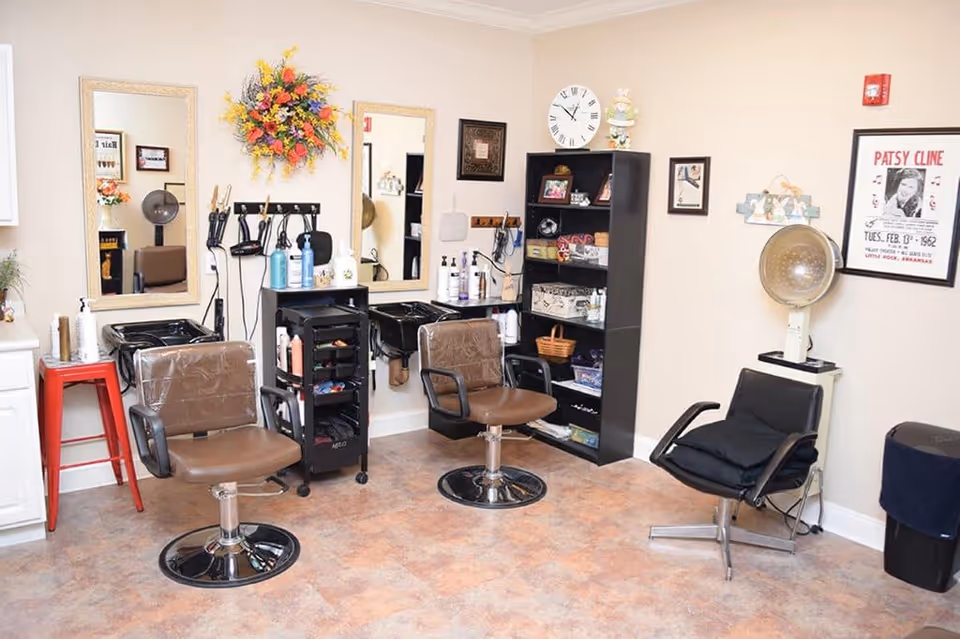 Interior of a hair salon area in a senior living facility with two brown salon chairs in front of mirrors, hair washing sinks, a black rolling cart with hair products, a black shelving unit with various items, and a vintage hair dryer chair. The walls are decorated with a floral arrangement and framed pictures.