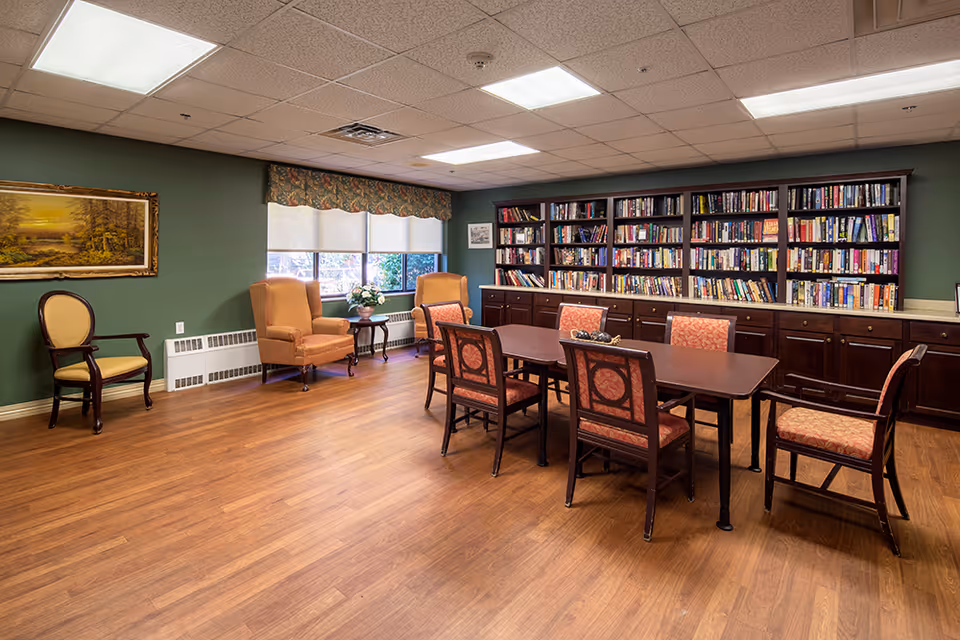 A cozy room with wooden flooring featuring a long table surrounded by six upholstered chairs. Along the back wall, there is a large bookshelf filled with books and cabinets underneath. Two armchairs and a small round table with a flower arrangement are placed near a window with white blinds and a floral valance. A framed landscape painting hangs on the green wall to the left.