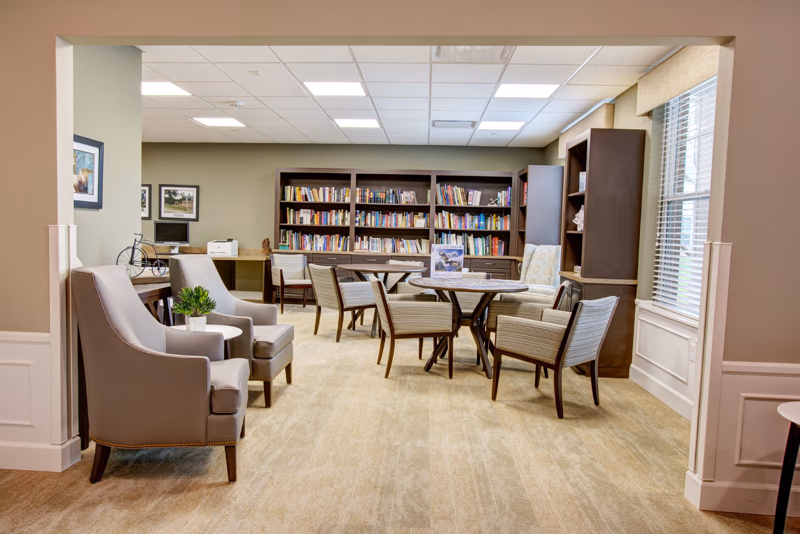 A bright communal library-style seating area with bookshelves, a round table surrounded by chairs, and upholstered armchairs by a window.