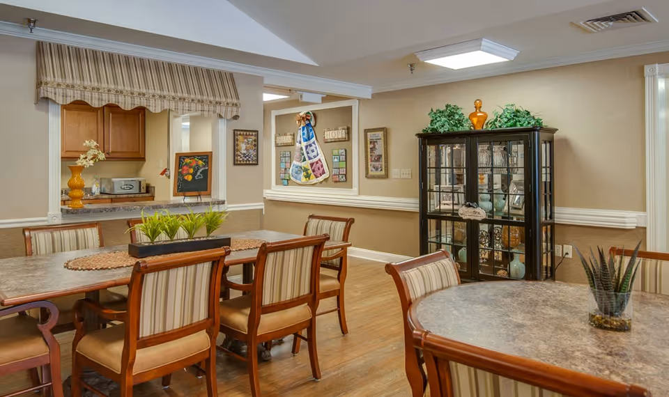 A cozy dining area with two tables surrounded by wooden chairs with striped cushions. The room features a display cabinet filled with decorative items, a small kitchen pass-through window with a striped valance, and wall decorations including framed pictures and a colorful quilt hanging. The floor is wooden, and the walls are painted beige with white trim.