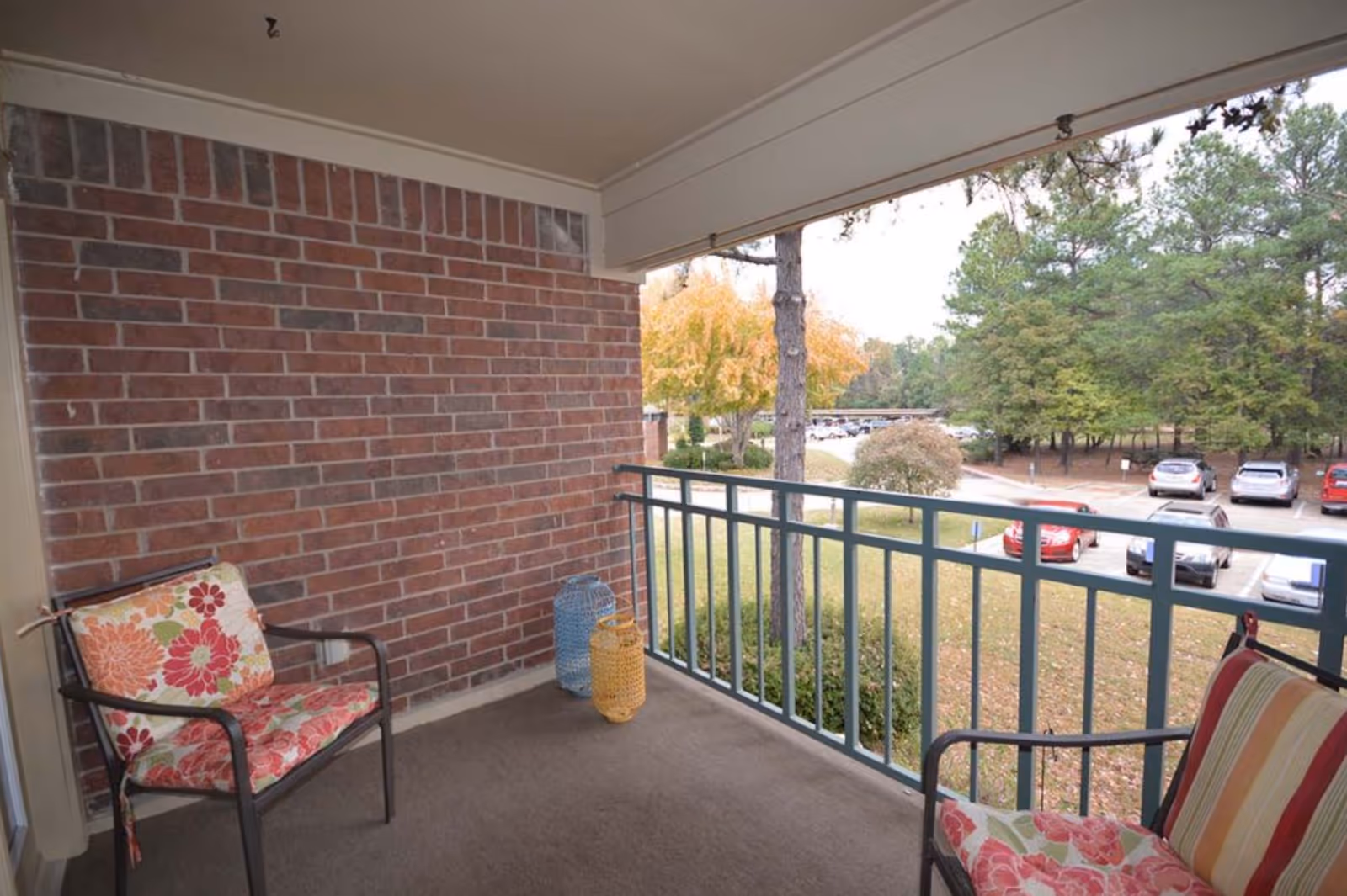 Covered apartment balcony with two patterned chairs, a brick wall, railing, and a view of a parking lot and trees.