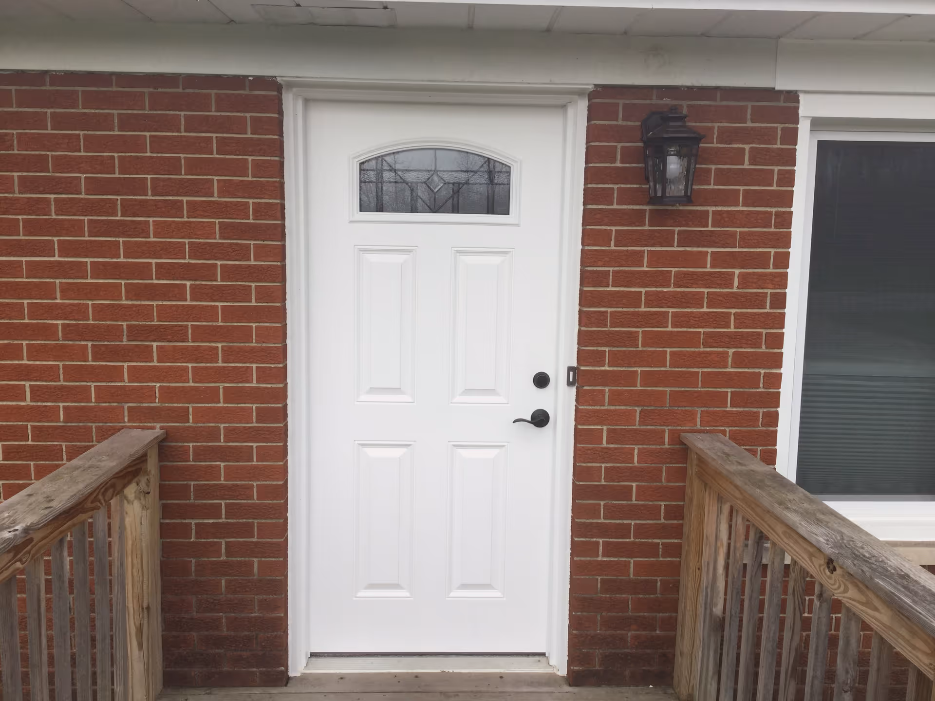 White entry door with a small arched window set in a red brick wall, flanked by wooden railings and an exterior light.
