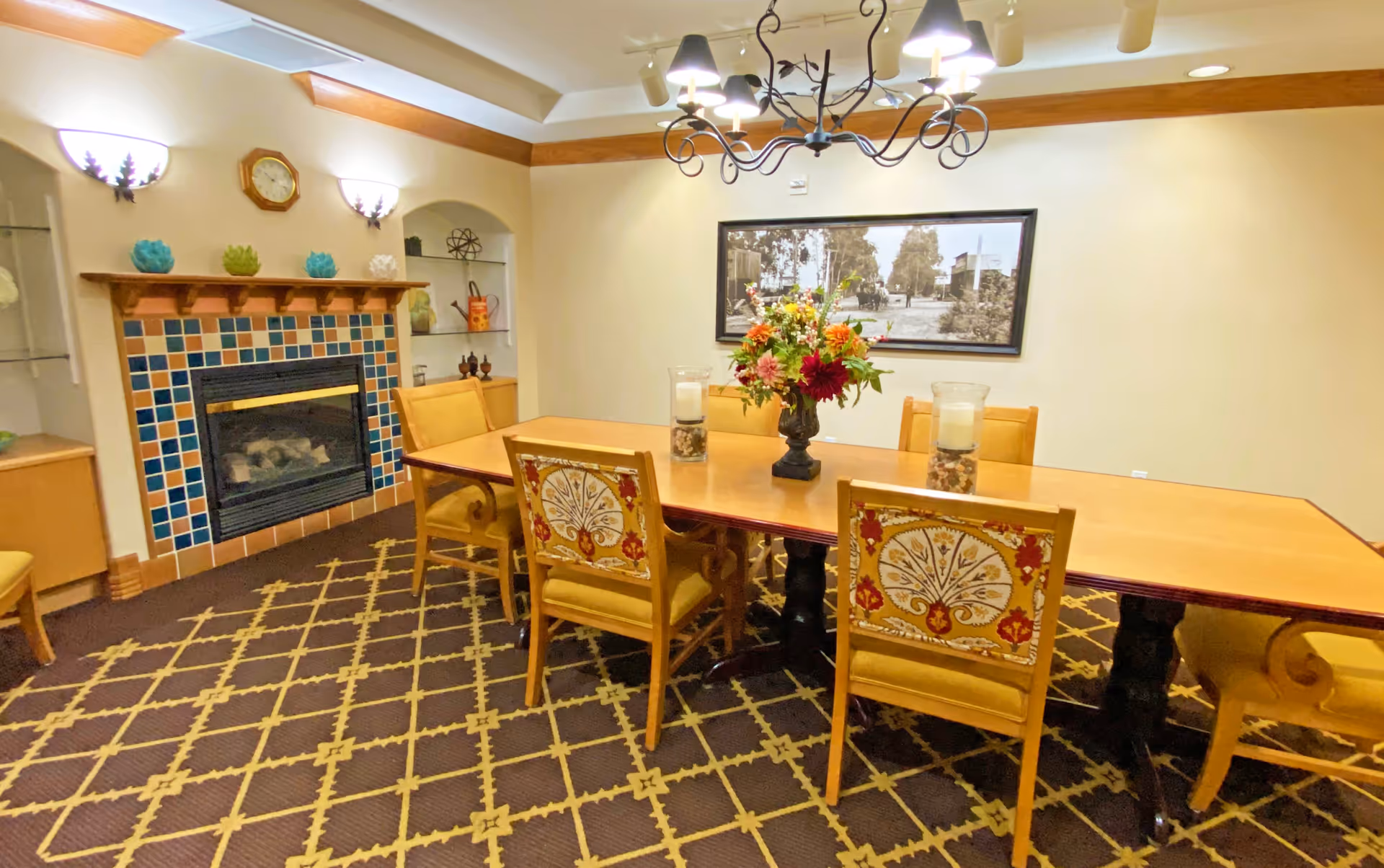 A dining room with a long wooden table surrounded by yellow upholstered chairs, some with decorative backs. The room features a fireplace with a colorful tiled surround, a patterned carpet, a chandelier hanging from the ceiling, and a framed black and white photograph on the wall. There are decorative items on shelves and a floral centerpiece on the table.